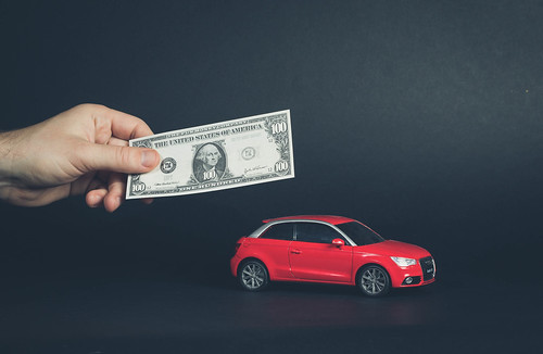 hand holding a 100 dollar bill in front of a miniature red car